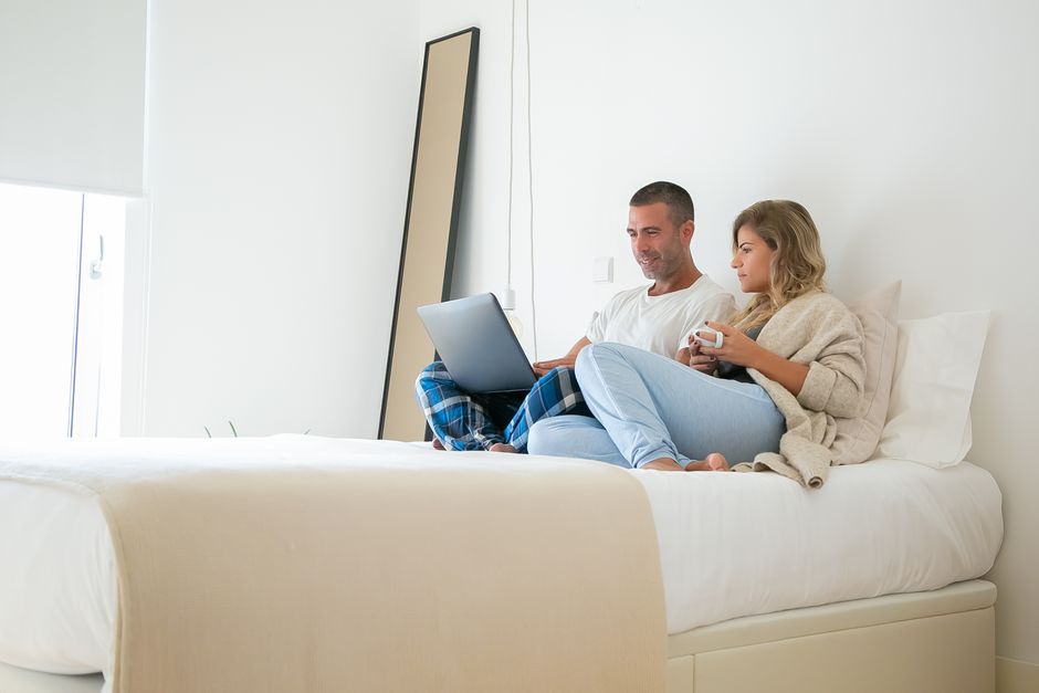 A man and woman relax on a bed, enjoying coffee and using a laptop in a bright bedroom setting.