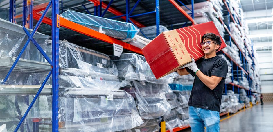 Smiling warehouse worker carrying a box in a large storage aisle filled with packaged goods.
